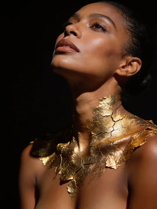 Woman with cracked gold leaf painted across her shoulders against a black backdrop